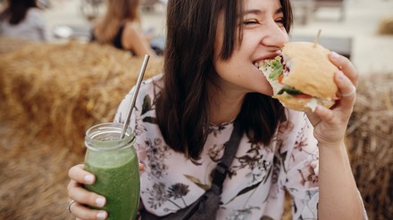 Frau isst einen Burger und hält einen grünen Smoothie in der Hand, draußen in einem Gastronomie-Umfeld.