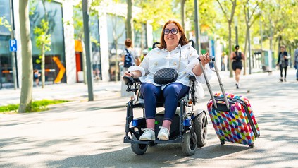 Frau im Elektrorollstuhl zieht einen Rollkoffer auf einer barrierefrei gestalteten Straße mit Passanten im Hintergrund.