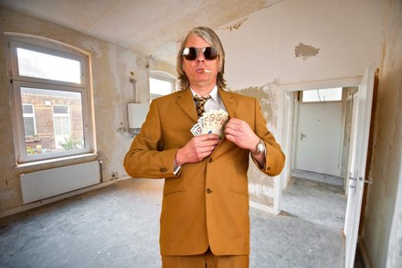 Ein Mann im Anzug mit Sonnenbrille und Zigarette steht in einem heruntergekommenen Gebäude und steckt sich mehrere Geldscheine in die Tasche