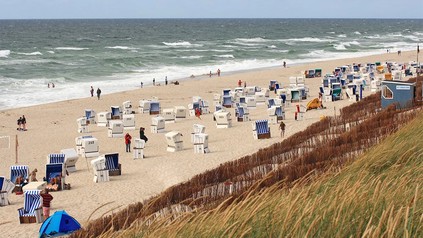 Strand an der Nordseeküste mit zahlreichen Strandkörben, Spaziergängern und rauer See unter bewölktem Himmel.