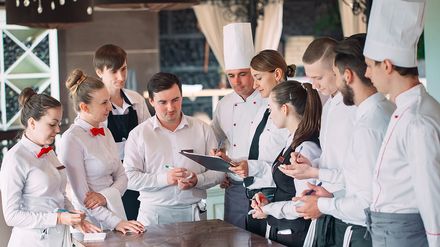 Hotel- und Gastronomieteam mit Küchenchef und Service-Mitarbeitern bei einer Teambesprechung im Restaurant.