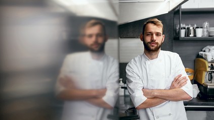 Junger Koch lehnt entspannt an einer Wand und lächelt im Arbeitsumfeld Küche