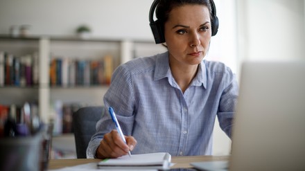 Frau mit Kopfhörer vor dem Laptop, schreibt Notizen auf