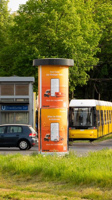 Lalamove-Werbesäule an einer Straße in Berlin mit vorbeifahrender Straßenbahn.