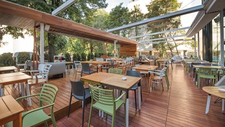 Holzterrasse im Außenbereich einer Gastronomie mit grünen Stühlen, Holztischen und einer geöffneten Terrassenüberdachung