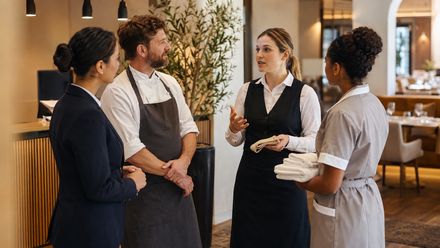 Vier Mitarbeiter aus Rezeption, Küche, Service und Housekeeping stimmen sich in einem Hotelrestaurant vor der Schicht ab.