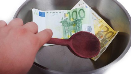 Eine Hand rührt mit einem Kochlöffel in einer Schüssel in der sich Geldscheine befinden