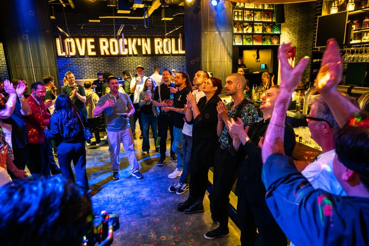 Zahlreiche Menschen stehen in einer Bar und applaudieren; im Hintergrund leuchtet ein „Love Rock ’n’ Roll“-Schriftzug.