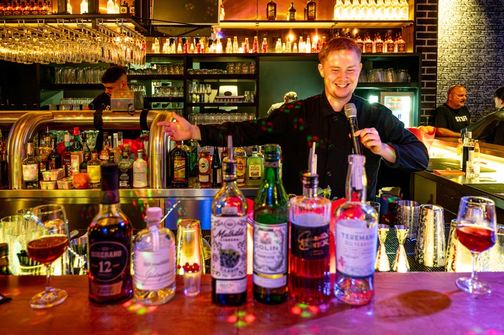 Ein lächelnder Bartender spricht hinter einer Bar in ein Mikrofon; davor stehen zahlreiche Flaschen und Gläser.
