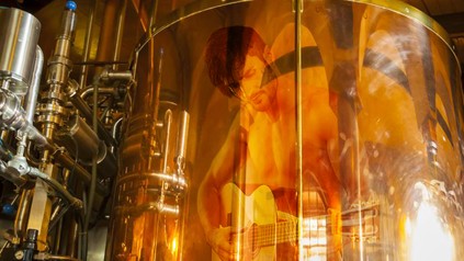 Ein großer Braukessel auf dem sich ein oberkörperfreier Mann mit Gitarre spiegelt