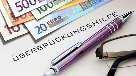 Euro-Geldscheine, ein Kugelschreiber neben einer Brille und das Wort Überbrückungshilfe