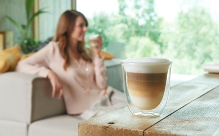 Ein modernes Glas mit Kaffee gefüllt und eine Dame im Hintergrund, die gerade aus einem solchen Glas trinkt