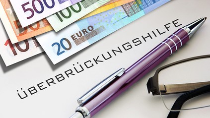 Euro-Geldscheine, ein violetter Kugelschreiber, eine Lesebrille und das Wort "Überbrückungshilfe"