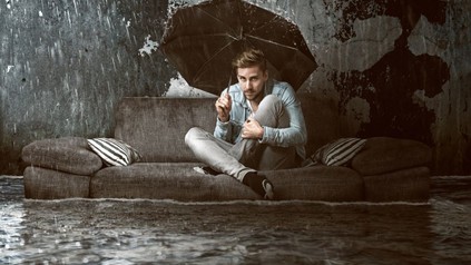 Mann unter Regenschirm auf Sofa in überfluteter Wohnung