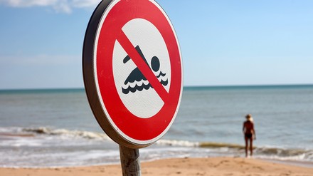 Strand mit einem „Schwimmen verboten“-Schild, im Hintergrund steht eine Person