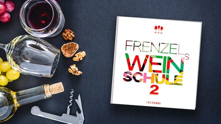 Das Buch Frenzels Weinschule 2 liegt neben einem Glas mit Rotwein, einem leeren Weinglas und einer Flasche Weißwein, Trauben, Nüssen und einem Flaschenöffner