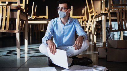 Frustrierter Gastronom mit Mund-Nasen-Maske sitzt auf dem Boden seines geschlossenen Restaurants, um ihn herum liegen Papierdokumente, die Stühle stehen umgedreht auf den Tischen