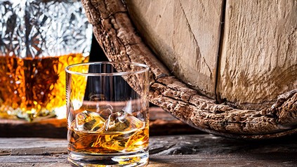 Ein Glas mit Whiskey und Eiswürfeln vor einem Holzfass