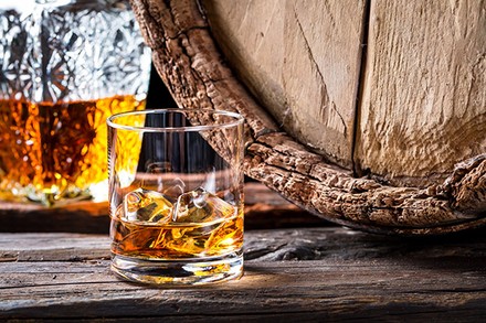 Ein Glas mit Whiskey und Eiswürfeln vor einem Holzfass