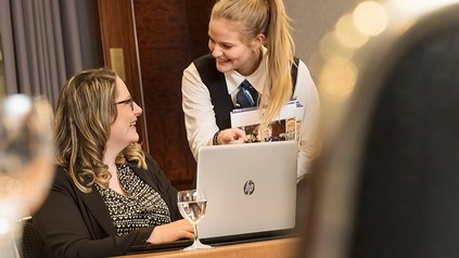 Eine Hotelmitarbeiterin spricht lächelnd mit einer Besucherin an der Rezeption vor einem Laptop