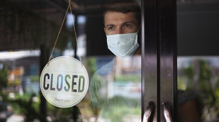 Ein Mann, Gastronom, steht mit einer Mund-Nasen-Maske hinter einer geschlossenen Glastür und blickt wartend nach draußen; an der Tür hängt ein Schild „Closed“, das Restaurant/Café ist geschlossen