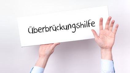 Ein untergehender Unternehmer hält ein Schild hoch, auf dem "Überbrückungshilfe" steht