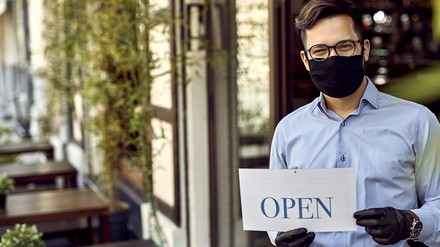 Kellner mit Maske und Handschuhen steht vor den Tischen eines Cafés oder Restaurants und hält ein Schild mit dem Wort „Open“
