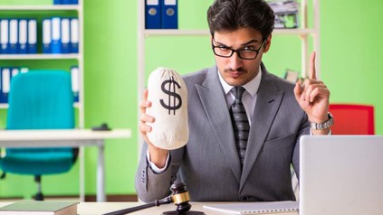 Ein Beamter vor einem Computer hält einen Sack Geld in der Hand und hebt mahnend den Finger