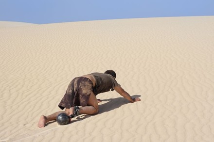 Mann in Lumpen robbt mit Fußkugel an Kette durch die Wüste