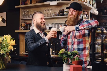 Zwei bärtige Männer, die sich hinter einer Restauranttheke mit Kaffeetassen zuprosten