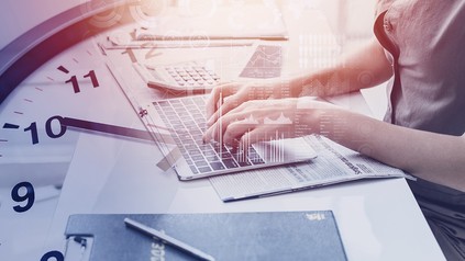 Nahaufnahme von Händen einer jungen Frau, die etwas auf einem Laptop tippt; das Bild wird von einer Uhr überlagert