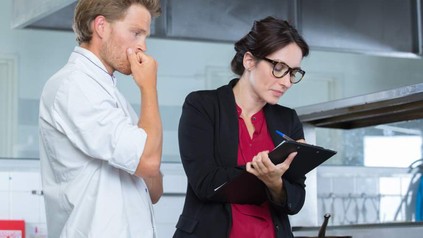 Frau notiert sich etwas auf Klemmbrett, Koch sieht besorgt zu