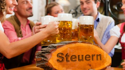 Eine Gruppe bayerisch angezogener Leute an einem Wirtshausstammtisch. Auf dem Stisch steht ein Holzschild, auf dem das Wort Steuern eingraviert ist.