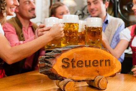 Eine Gruppe bayerisch angezogener Leute an einem Wirtshausstammtisch. Auf dem Stisch steht ein Holzschild, auf dem das Wort Steuern eingraviert ist.