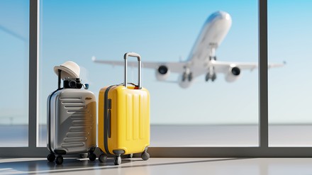 zwei Koffer stehen am Flughafen vor einer Glaswand, im Hintergrund startet ein Flugzeug