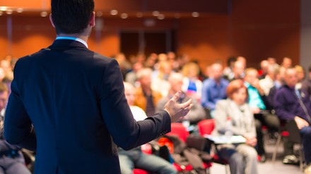 Mann steht auf der Bühne vor vollem Tagungssaal