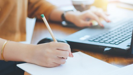 Nahaufnahme einer jungen Frau, die an einem Laptop sitzt und etwas auf einen Block schreibt