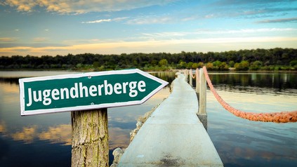 Ein Pfeilschild mit dem Wort „Jugendherberge“ zeigt auf einen Steg, der über einen See führt