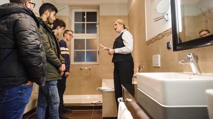 Hotel-Mitarbeiterin erklärt Jugendlichen die Reinigungsarbeit in einem Badezimmer
